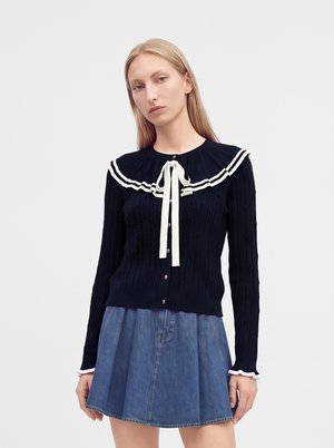 Jeune femme portant un haut noir avec un nœud blanc et un col, une jupe plissée bleue, des chaussettes blanches, des chaussures blanches, tenant un petit sac marron.