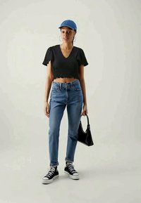 Top curto de manga curta preto, jeans azuis de cintura alta, tênis pretos com solas brancas, e um boné azul, segurando uma bolsa preta.