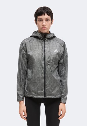 Personne portant une veste à capuche grise avec un petit logo sur la poitrine et un pantalon noir, debout face à l'avant sur un fond uni.