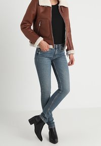 Giacca di pelle marrone con colletto in pile, maglietta nera e jeans skinny blu. Stivaletti neri alla caviglia completano l'outfit.