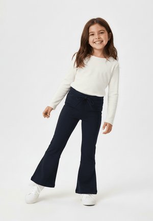 Uśmiechnięta dziewczyna o długich brązowych włosach, ubrana w biały top z długim rękawem, granatowe rozszerzane spodnie i białe trampki na białym tle.