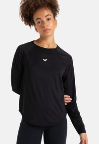 Nuori nainen, jolla on kiharat hiukset, pukeutuneena mustaan pitkähihaiseen urheilupaitaan, jossa on pieni valkoinen logo rinnassa, poseeraa käsi selän takana.