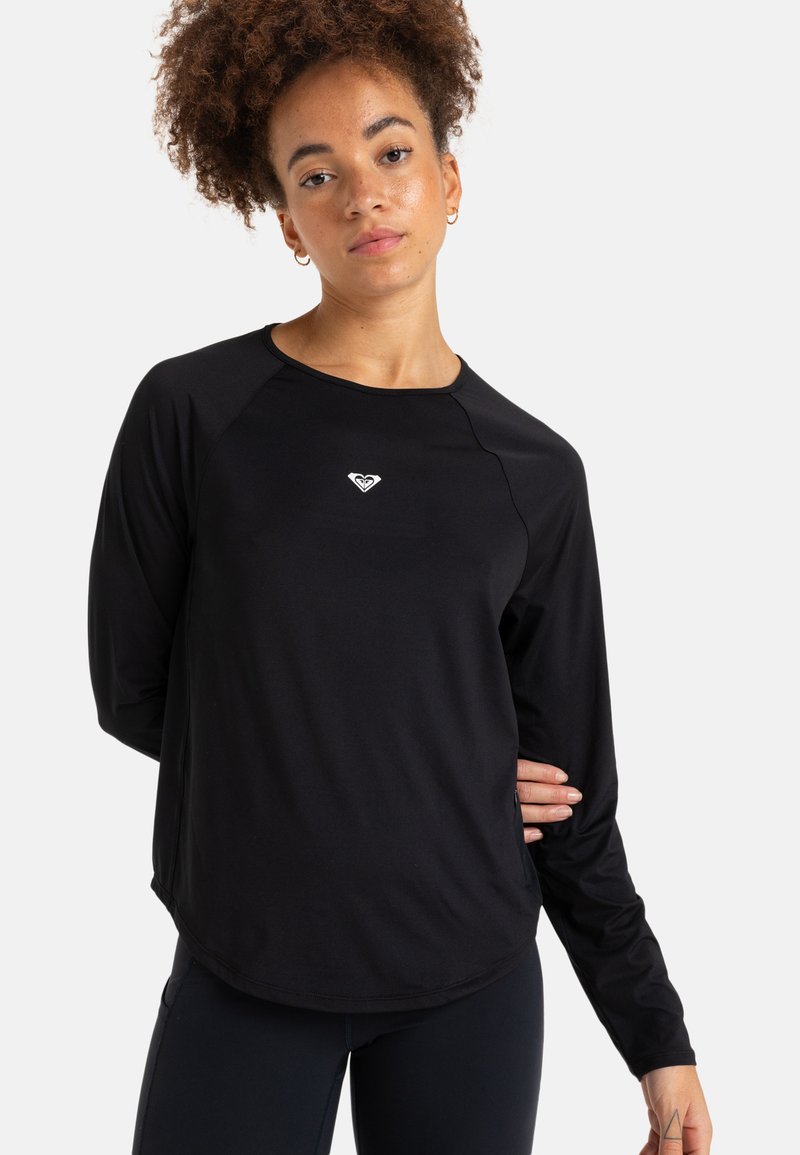 Nuori nainen, jolla on kiharat hiukset, pukeutuneena mustaan pitkähihaiseen urheilupaitaan, jossa on pieni valkoinen logo rinnassa, poseeraa käsi selän takana.