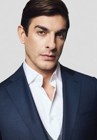 Granatowy garnitur z teksturowanym wzorem, w zestawieniu z białą koszulą i szarym kamizelką. Gładkie włosy, neutralne tło. Portret z bliska.