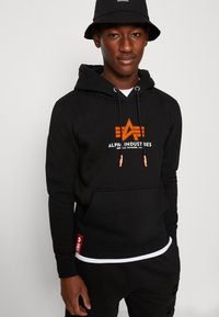 Sudadera negra de mezcla de algodón, con un bolsillo frontal, logo naranja y detalles en naranja en los cordones. Etiqueta roja en el dobladillo.