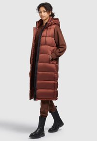 Brauner Puffercoat mit langem Design, Kapuze und sichtbarem Nähen; kombiniert mit braunen Hosen und schwarzen Stiefeln. Glatte, glänzende Stoffoberfläche.