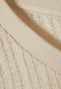 Strikkegenser i lys beige med teksturert, åpen vevmønster. Har en ribbestrikket halskant og glatt stofffôr. Myk, luftig utseende.