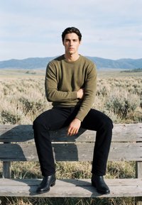 Jeune homme en pull vert et pantalon noir assis sur un banc en bois en plein air avec des montagnes et de l'herbe sèche en arrière-plan.