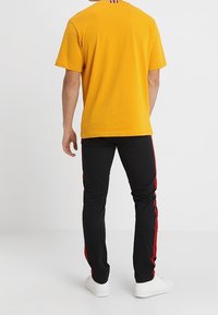Gul kortärmad t-shirt med rund halsringning, kombinerad med svarta slim fit-byxor med röda sidostripes och vita sneakers.
