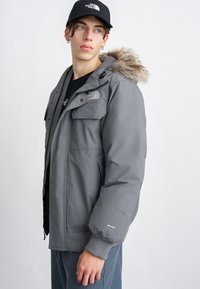 Grau Outdoor-Jacke mit einer Kapuze aus Kunstfell, Reißverschluss vorne und elastischen Bündchen; sie verfügt über mehrere Taschen und markenspezifische Logo-Details.