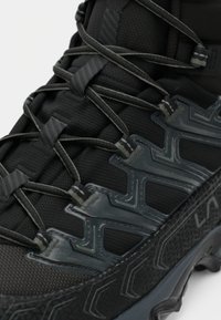 Close-up de uma bota de trekking preta com um atacador detalhado, parte superior em tecido texturizado e borda de sola estampada.