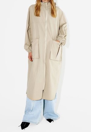 Personne portant un long manteau beige à capuche avec fermeture éclair, grandes poches avant, sur un jean large bleu clair et des chaussures noires pointues.