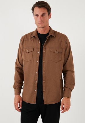Bruine overhemd met knopen in een zachte stof, voorzien van twee borstzakken, drukknoopsluiting en een klassieke kraag. Draagt over een zwart t-shirt.