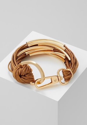 Bracelet en cordon marron tressé avec des tubes métalliques dorés incurvés, fermoir doré et anneau, présenté sur une surface blanche inclinée.