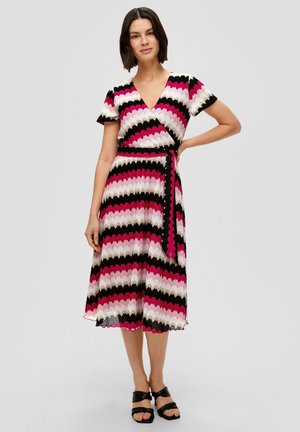 Wikkeljurk met een zigzagpatroon in roze, zwart en gebroken wit, met korte mouwen, een strik taille en een lengte tot de knie. Zwarte sandalen.