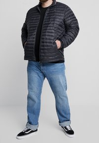 Svart kviltad puffjacka med ståkrage, dragkedja fram och två sidofickor, parat med ljusblå jeans och svarta sneakers.