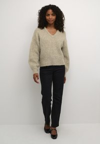 Pull en tricot beige avec un col en V et des épaules tombantes, associé à un pantalon noir à jambe droite et des chaussures noires. Texture plate et coupe décontractée.