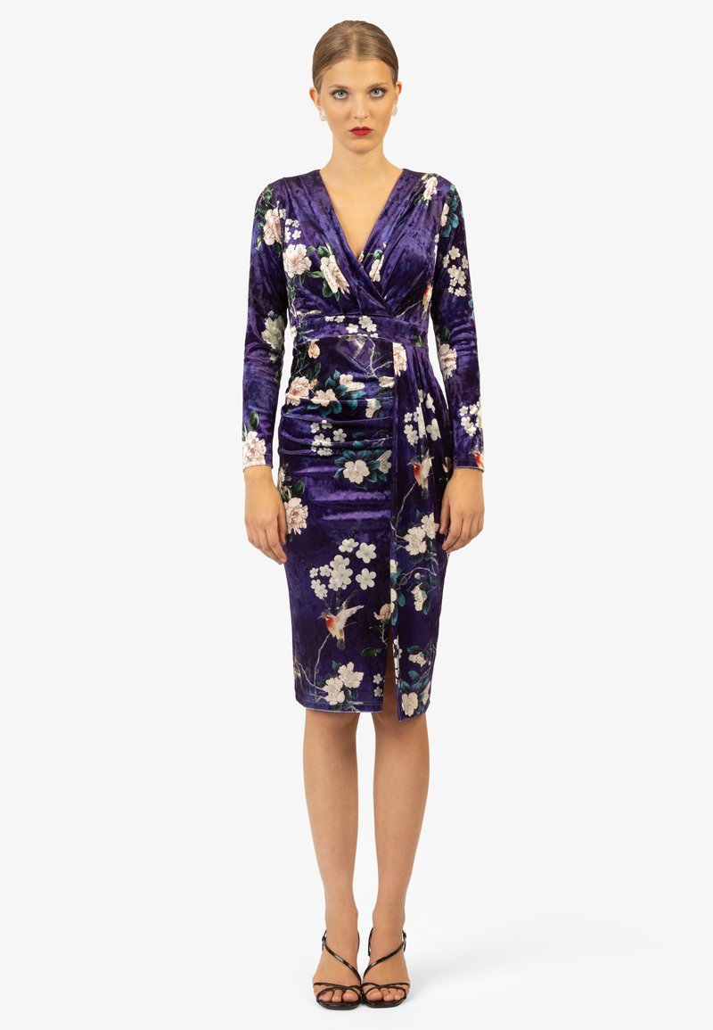 Paarse fluwelen jurk met lange mouwen, bloemenpatronen, V-hals en een aansluitende pasvorm. Heeft een zijsplit en een wikkeldesign.