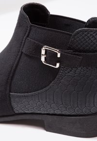 Botín negro hasta el tobillo con una sección de cuero texturizado con patrón de reptil y un detalle de hebilla metálica en la banda elástica. Parte superior de tela suave.