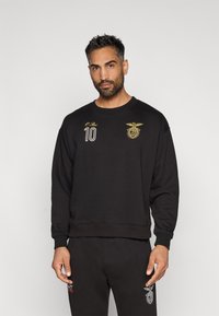 Uomo con capelli corti e scuri e barba, che indossa una felpa nera e pantaloni neri con un logo sportivo dorato e bianco e il numero 10 sulla parte anteriore della felpa.