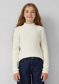 Junges Mädchen mit langen Haaren, das einen weißen strukturierten Pullover und dunkelblaue Jeans trägt, steht vor einem schlichten hellgrauen Hintergrund.