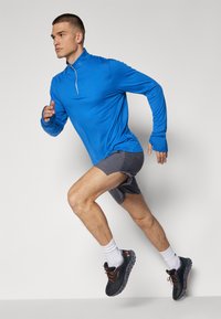 Sportlik riietus, kus on sinine pikkade varrukatega särk ja hallid lühikesed püksid. Mustad jooksujalatsid oranžide aktsentidega. Mudel jooksupositsioonis.