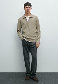 Ung man står klädd i en beigefärgad långärmad zip-neck tröja, en vit undertröja, mörkgrå jeans och bruna mockaskor på ett grått golv.