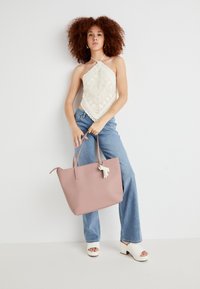 Sac fourre-tout en cuir rose avec de longues poignées, équipés d'un charm. Modèle portant un haut halter en crochet crème et un jean bleu clair. Sandales compensées blanches.