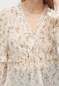 Blusa leggera e trasparente con stampa floreale in arancione e beige, con dettagli arricciati intorno al collo e alle maniche, e texture a pois.