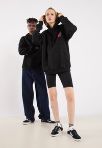 Black hooded sweatshirts with front pockets, worn over black cycling shorts and denim pants. Co-ordinated footwear features stripes.
