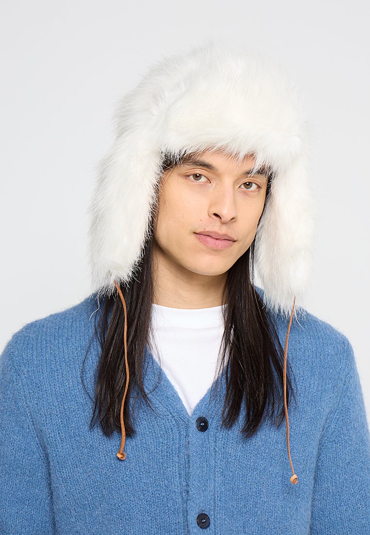 Erwachsene tragen einen weißen Kunstpelz-Trapperhut mit Ohrenschützern, eine blaue Strickjacke und ein weißes Hemd vor einem schlichten, hellen Hintergrund.