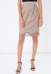 Falda beige a la altura de las rodillas con cintura ajustada y diseño plisado. El dobladillo presenta un borde de encaje con forma de concha, añadiendo textura y detalle.