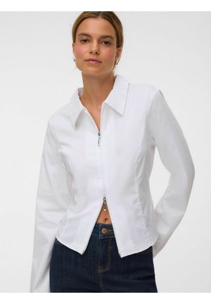 Kvinde iført en tætsiddende hvid langærmet lynlåstrøje og mørkeblå jeans, stående mod en ensfarvet lys baggrund.