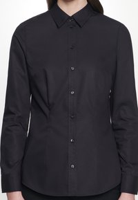 Kvinna iförd en åtsittande svart långärmad skjorta med knappar och klassisk krage, sedd från nacke till höfter mot en vit bakgrund.