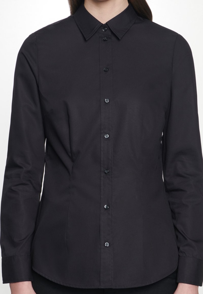 Kvinna iförd en åtsittande svart långärmad skjorta med knappar och klassisk krage, sedd från nacke till höfter mot en vit bakgrund.