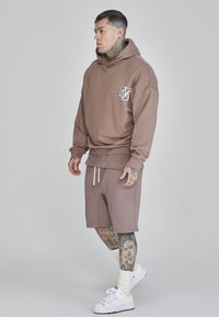 Sudadera marrón con bolsillo tipo canguro y logo, combinada con pantalones cortos a juego. Exhibida en una persona tatuada, que lleva zapatillas y calcetines blancos.