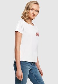 T-shirt en coton blanc à manches courtes et col rond, avec un design graphique rouge sur la poitrine. Porté avec un jean bleu.