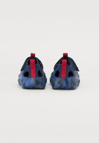 Zapatos azul marino con un patrón de camuflaje, parte superior de tela, lengüeta roja en el talón, diseño slip-on y perforaciones para ventilación.