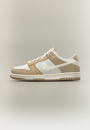 Baskets basses beige et blanches avec virgule Nike blanche, lacets blancs et semelle beige, présentées de profil sur un fond neutre.