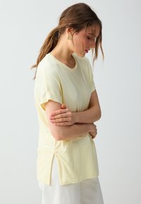 Femme aux cheveux bruns attachés en arrière, portant un t-shirt jaune pâle à manches courtes et un pantalon blanc, regardant vers le bas, les bras croisés.