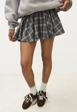 Personne portant une jupe plissée à carreaux grise, un sweat-shirt gris, des chaussettes blanches et des baskets marron foncé avec des lacets blancs sur un fond uni.