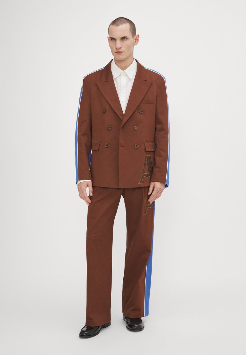 Giacca da uomo marrone doppiopetto con strisce laterali blu, pantaloni marroni coordinati con un logo in rilievo e camicia bianca sotto.