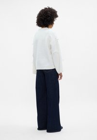 Baltas megztinis su raištelių akcentais ant rankovių, derinamas su laisvai krentančiomis tamsiai mėlynomis džinsinėmis kelnėmis. Lygi fono būsena.