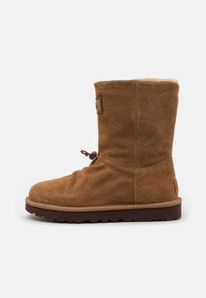 Botín de ante marrón hasta el tobillo con punta redonda y forro de piel, con un cierre lateral con toggle. Suela de goma con agarre texturizado y parte superior rematada.