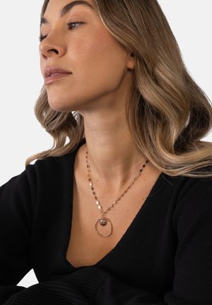 Frau mit hellbraunem, welligem Haar trägt eine runde goldene Halskette mit einem schwarzen Steinanhänger, bekleidet mit einem schwarzen V-Ausschnitt-Top.