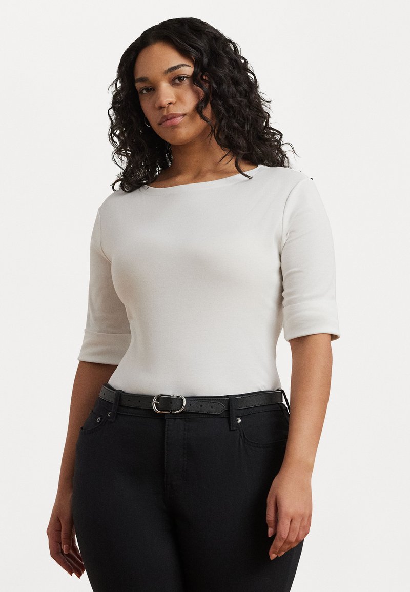 Top branco ajustado com decote round e mangas três quartos, combinado com jeans pretos de cintura alta e um cinto preto com fivela prateada.