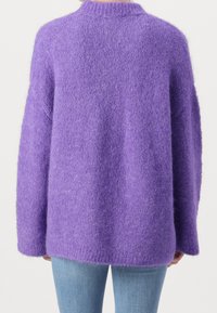 Kvinna i en mjuk, stor lila tröja och ljusblå jeans, sedd bakifrån mot en enkel bakgrund.