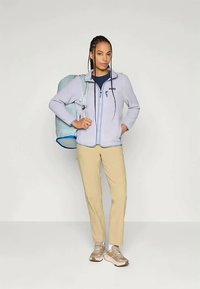 Heledsinine fleece'ist jakk, millel on ees lukku, beežid püksid ja heledat värvi pruunid tossud. Mudel hoiab käes sinist seljakotti, millel on sile tekstuur.