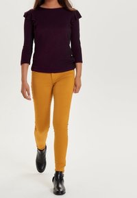 Tmavě fialový top s volánovými rukávy, přiléhavý střih. Hořčičně žluté skinny džíny. Černé kotníčkové boty. Jednoduché světlé pozadí.