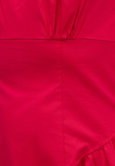 Κόκκινο ύφασμα με κάθετη ραφή και μαζεμένη λεπτομέρεια στις επάνω και κάτω άκρες, εμφανίζοντας υφή και ραφές.